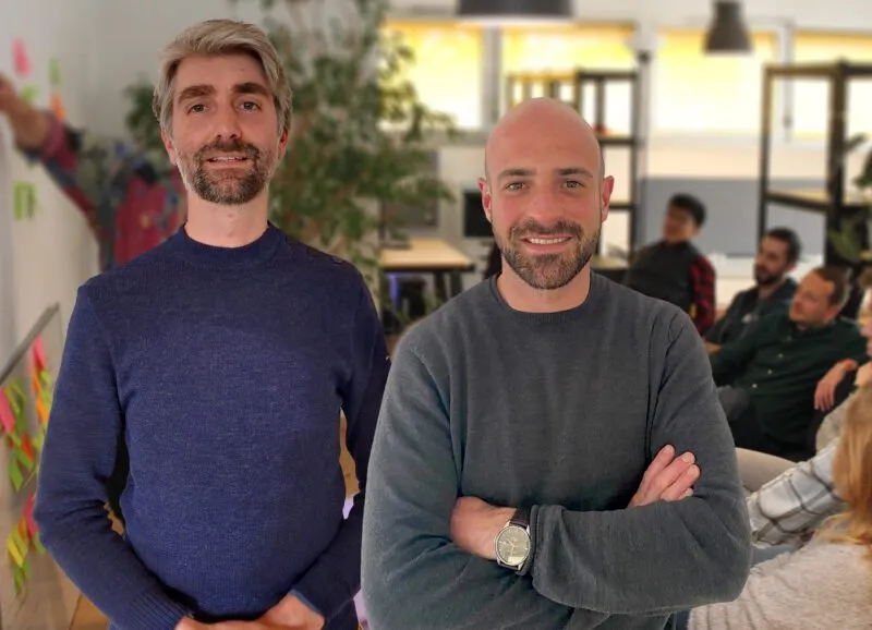 Two Picterra co-founders standing side by side indoors with arms crossed, with team members blurred in the background