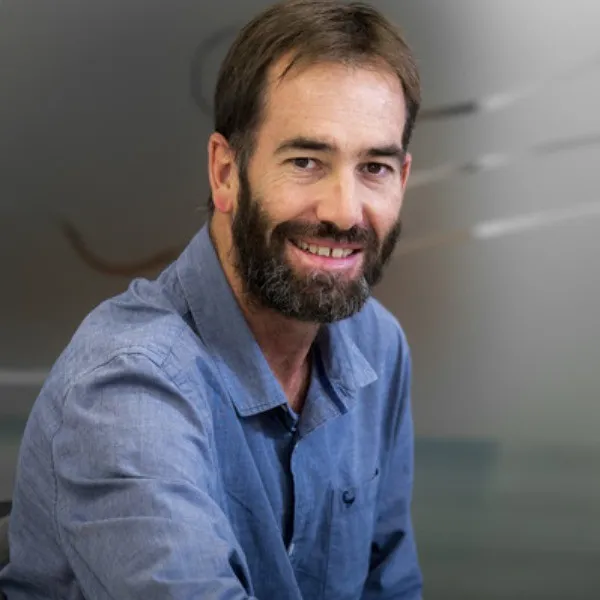 Portrait of a smiling man with a beard wearing a blue shirt, seated indoors against a softly blurred background.
